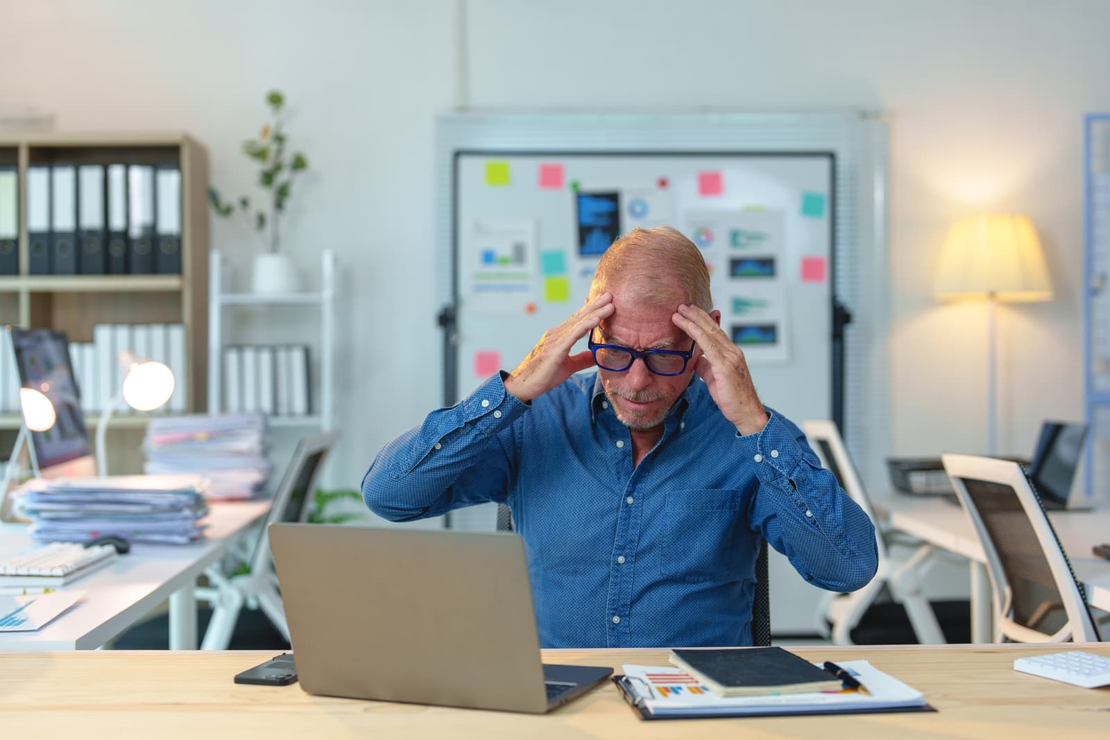 Senior manager struggling with a severe headache while working on a laptop in a chaotic office, massaging his