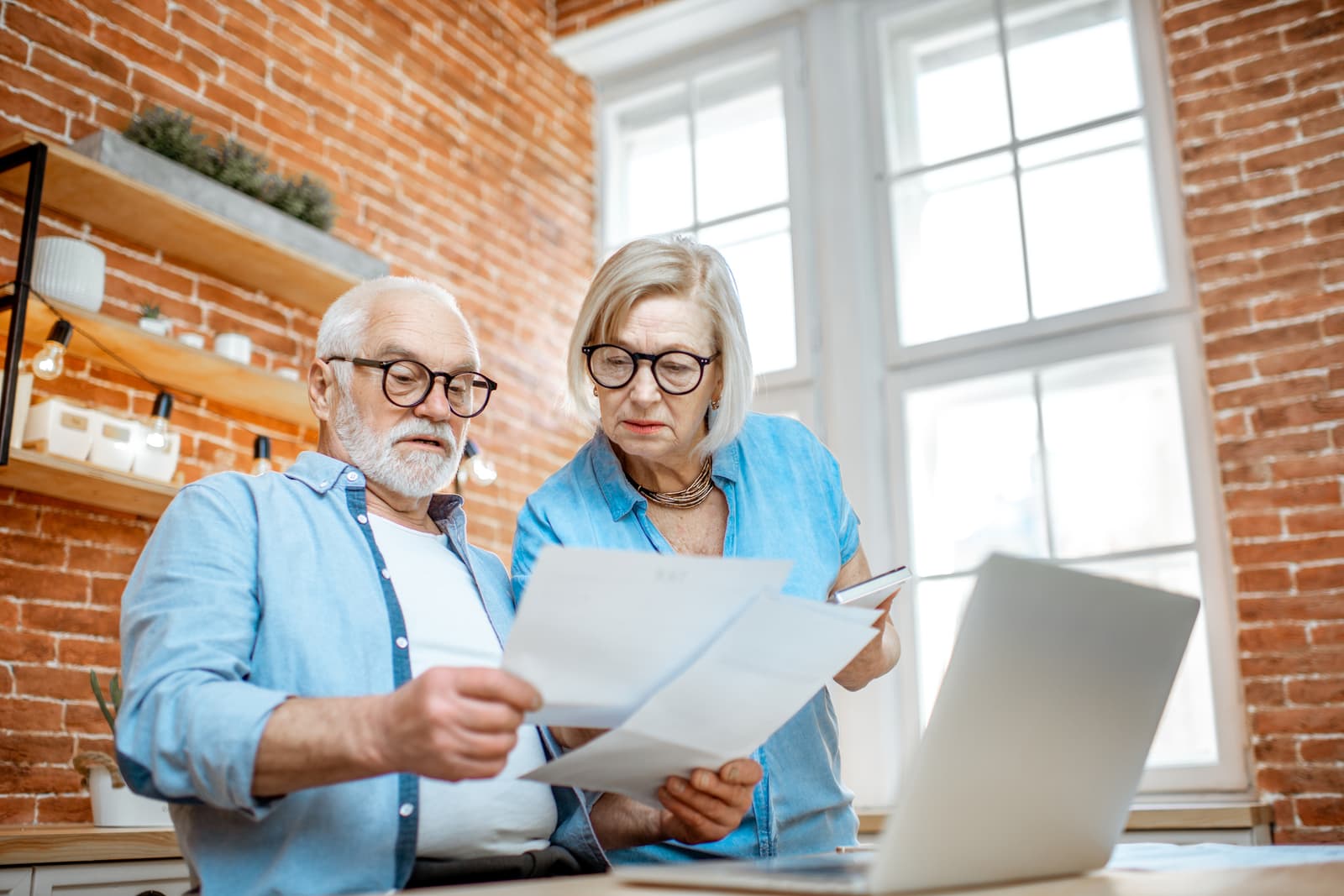 Senior couple with embarrassed emotions calculating some bills or taxes sitting with laptop on the kitchen at home