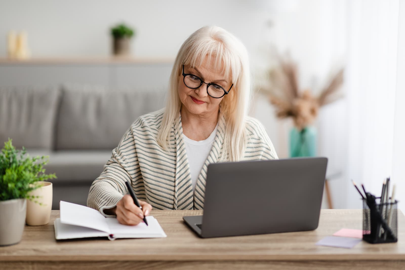 Mature lady writing in notebook and using laptop at home