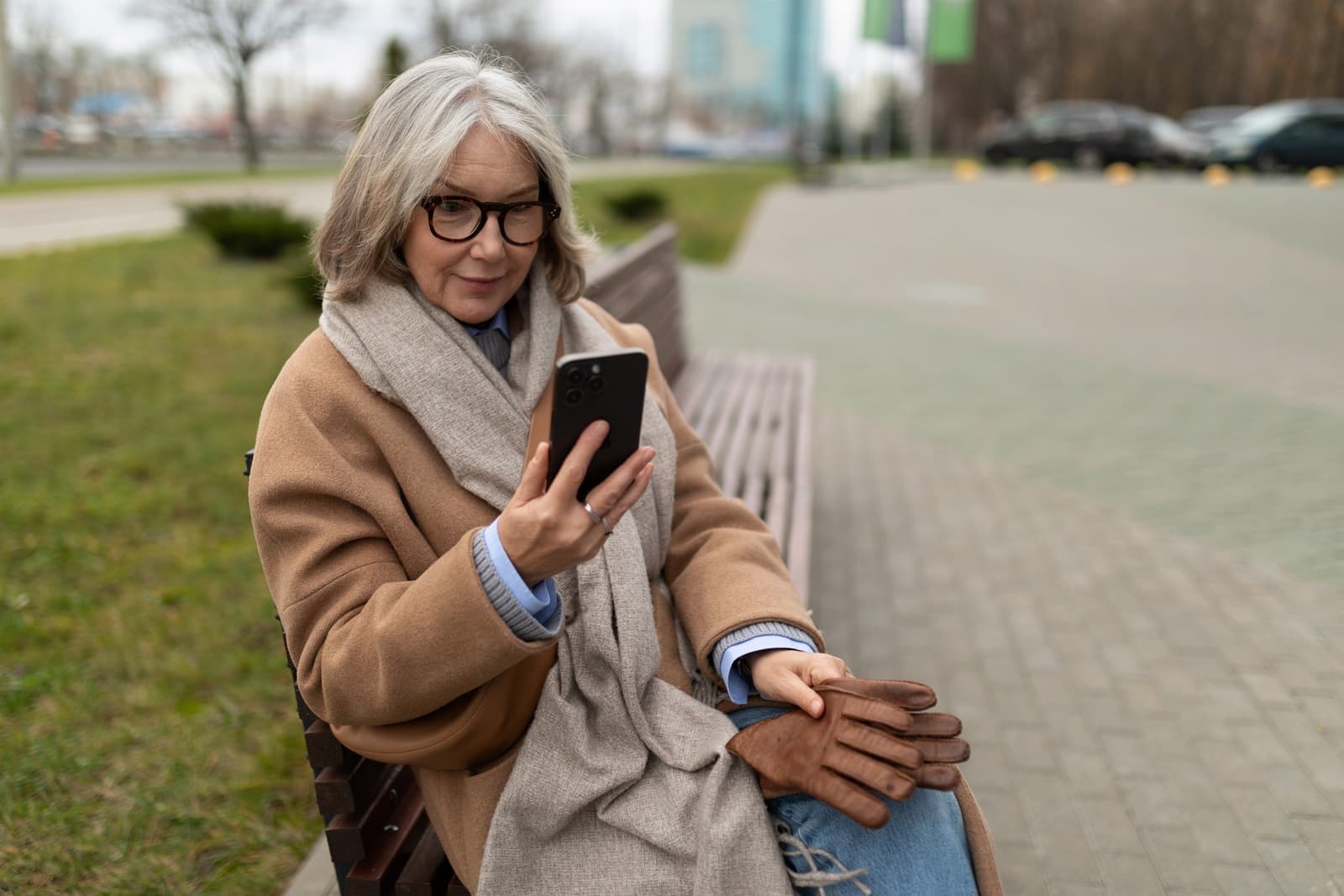 A grey-haired woman in glasses checking her phone on a park bench
