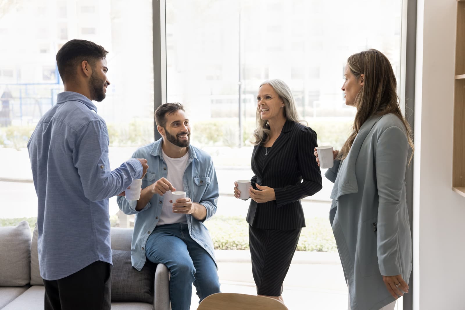 Group of four professionals enjoy coffee break in office environment, standing in workplace lobby holding cups,