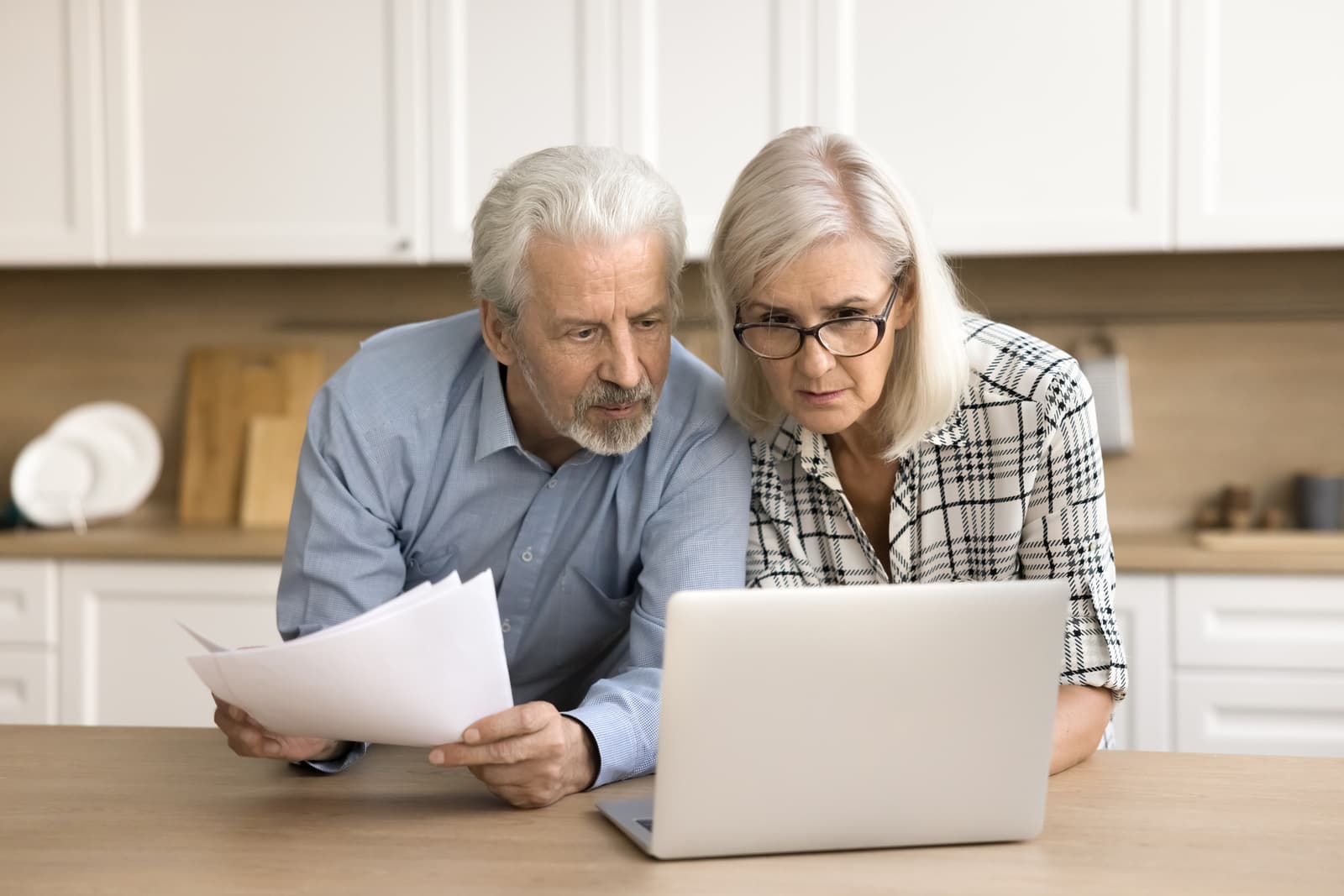 Serious senior couple checking domestic paper documents at kitchen table, using online financial banking application on